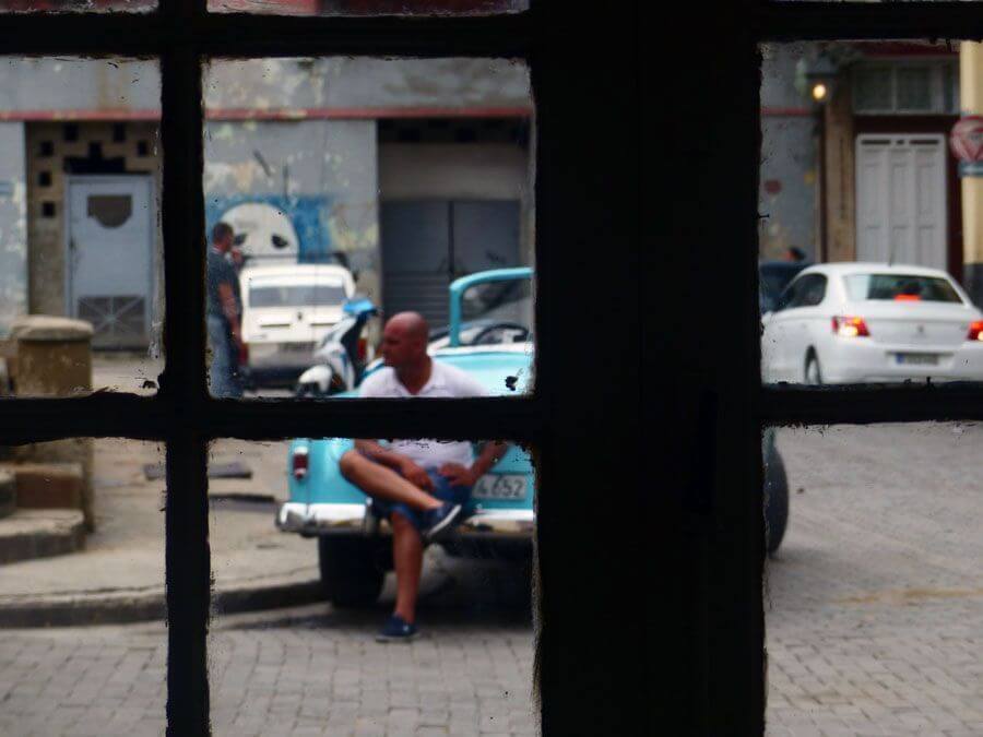 hombre cubano apoyado sobre su coche, fotografiado a través de una ventana